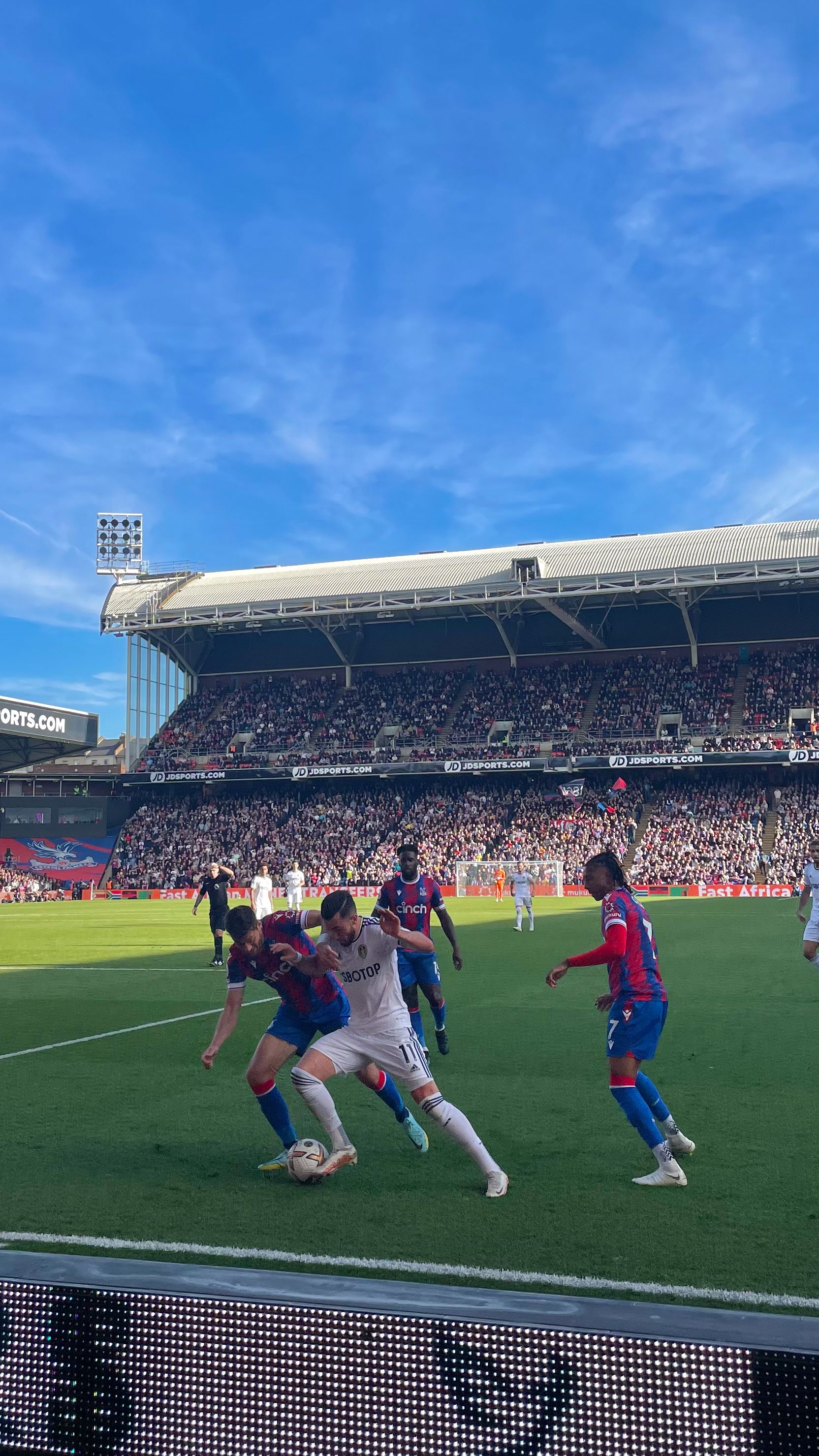 footballers battling for the ball with the stadium behind and a blue sky above