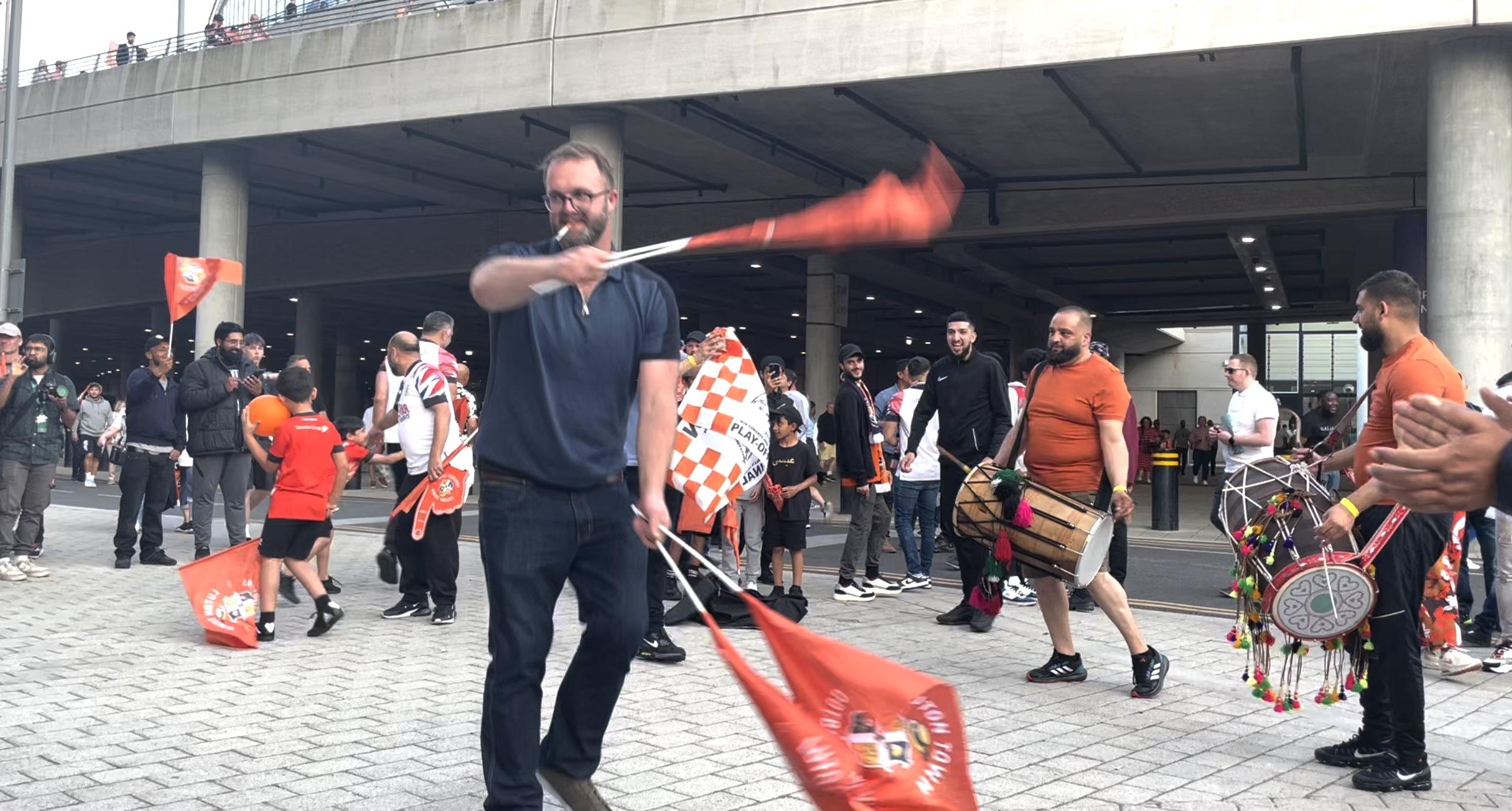 Man dancing with flags swirling in both hands, cigarette in mouth. Other men beat drums behind him.